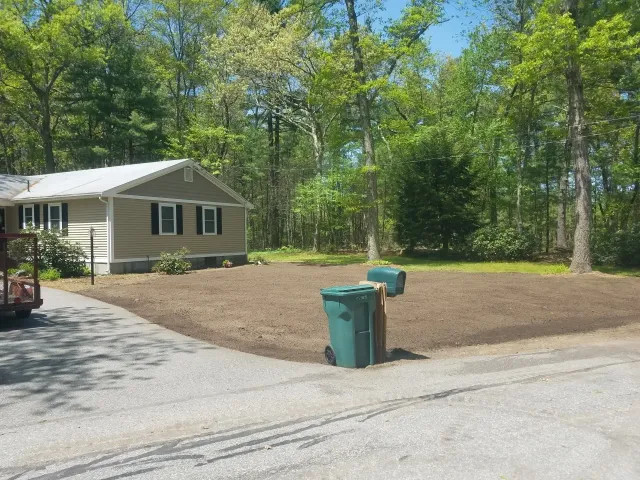 Small beige house by a gravel driveway with a dirt yard and green trees; two trash bins in front.