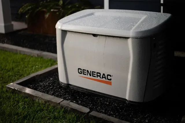Generator, gray casing, on gravel base, near brick wall. Gas pipes connected.