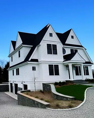 Completed custom-built New England home with white horizontal siding, black trim windows, multi-gabled roof, and gravel driveway.