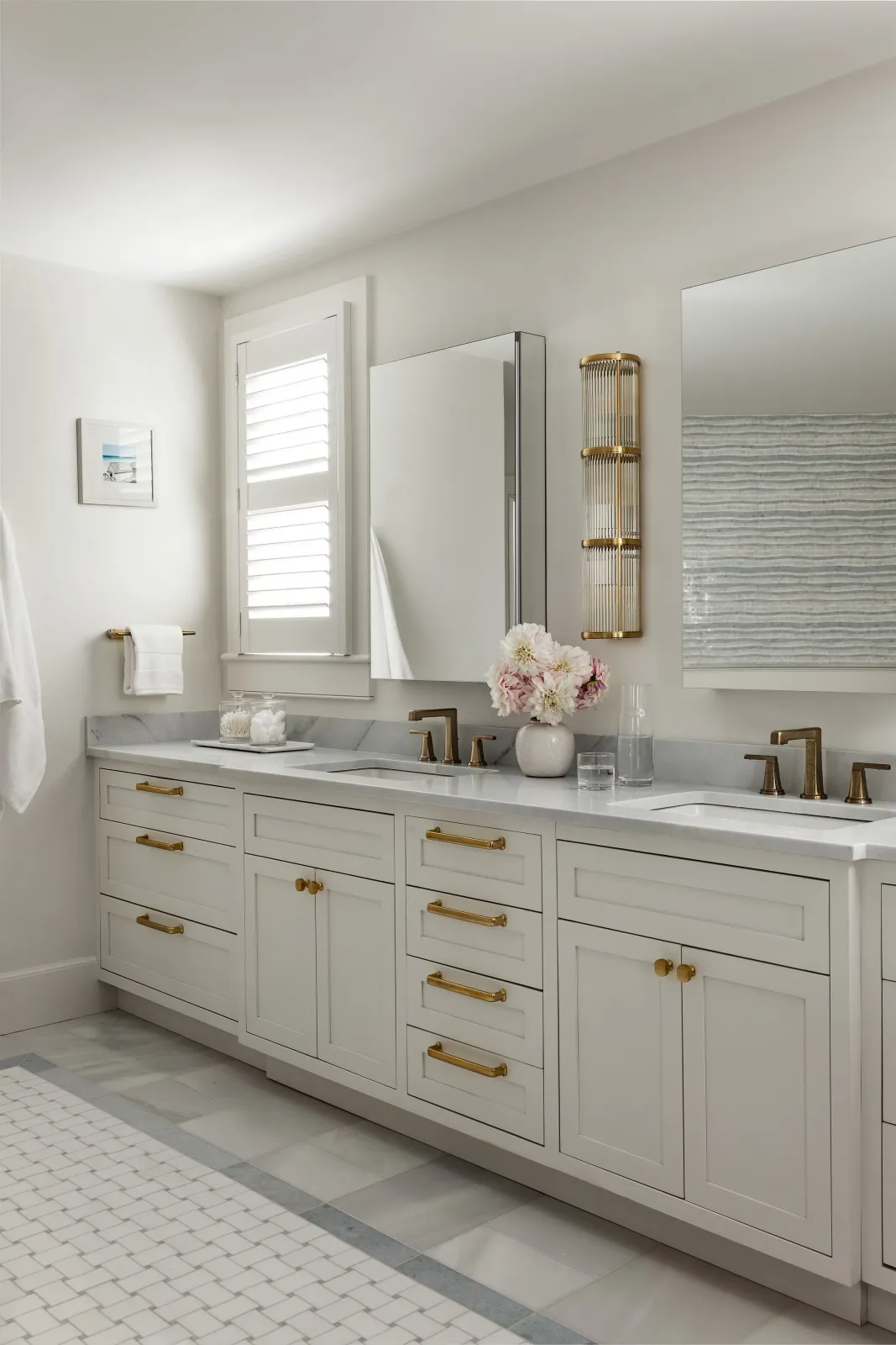 A modern white bathroom vanity with gold hardware, two sinks, and a marble countertop. There is a framed mirror, a floral vase, and a glass of water on the counter. Light enters through a small window with white shutters, and a towel hangs on a gold