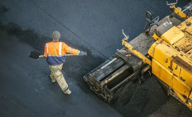 A worker in safety gear walks beside a yellow road paving machine on black asphalt.