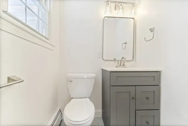 A half bathroom with white walls, a white toilet, and a grey vanity cabinet with a rectangular mirror and light fixture.
