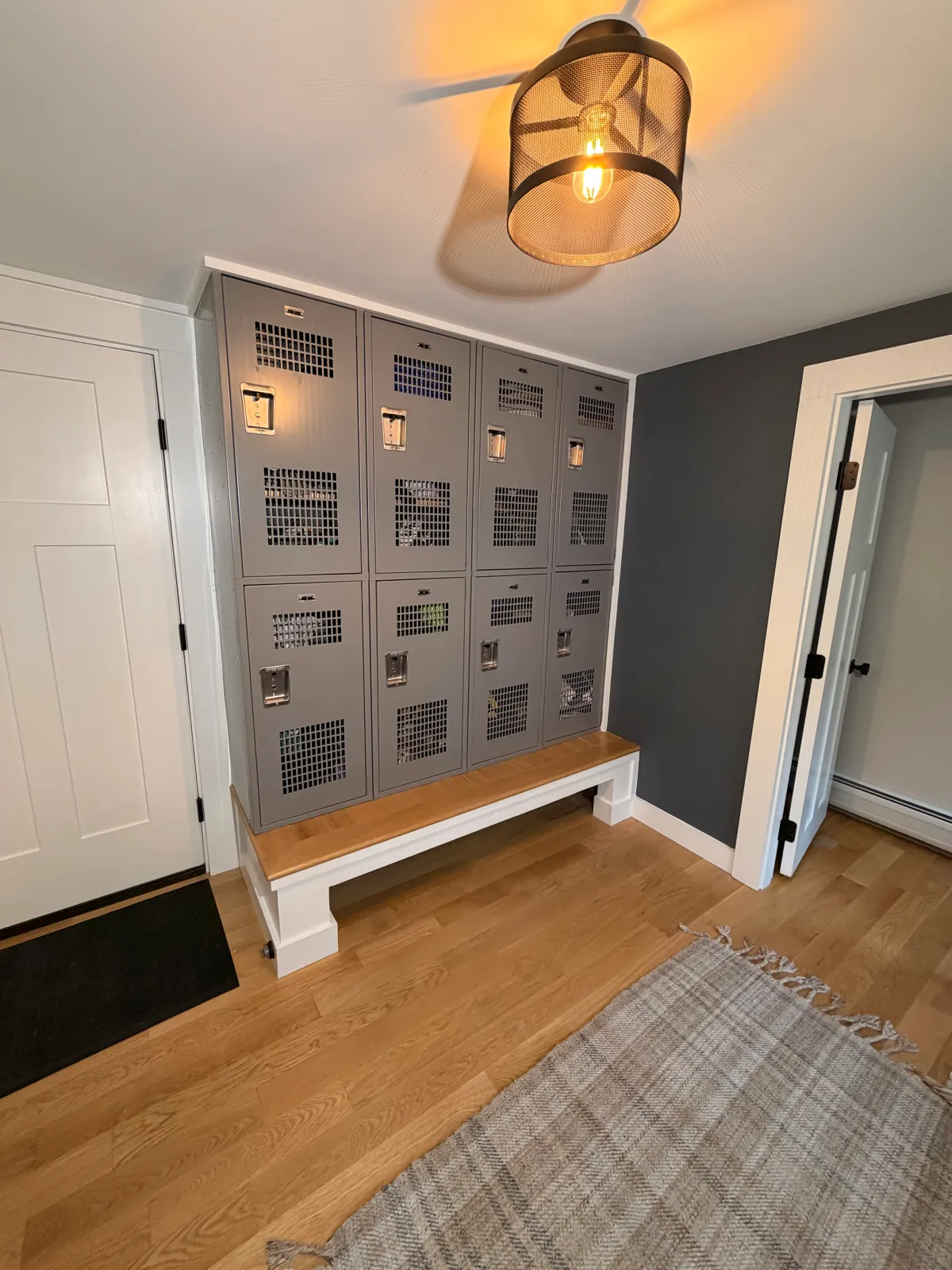 Gray metal lockers with ventilation holes, a handle on each door, and a rectangular lock. They are placed against a wall with a dark gray accent and sitting on a wooden bench with a white frame. Nearby is a green mat and a beige woven rug on a hardwood floor. The room has a ceiling mounted black cage light fixture with a visible filament bulb and white trim around the door frames.