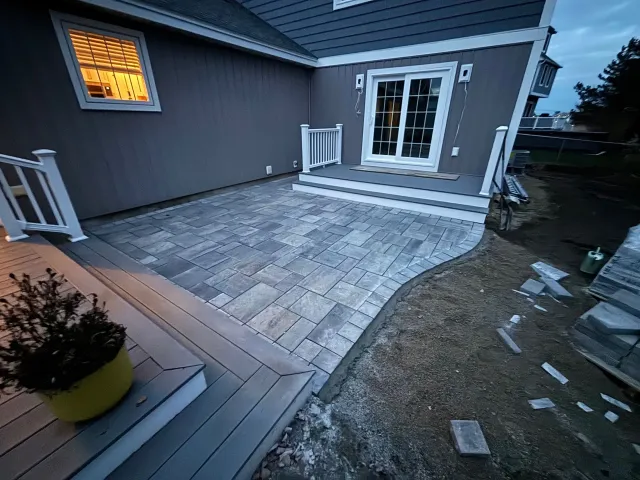 Gray brick patio next to a gray composite deck, leading to a sliding door. Exterior of a house.