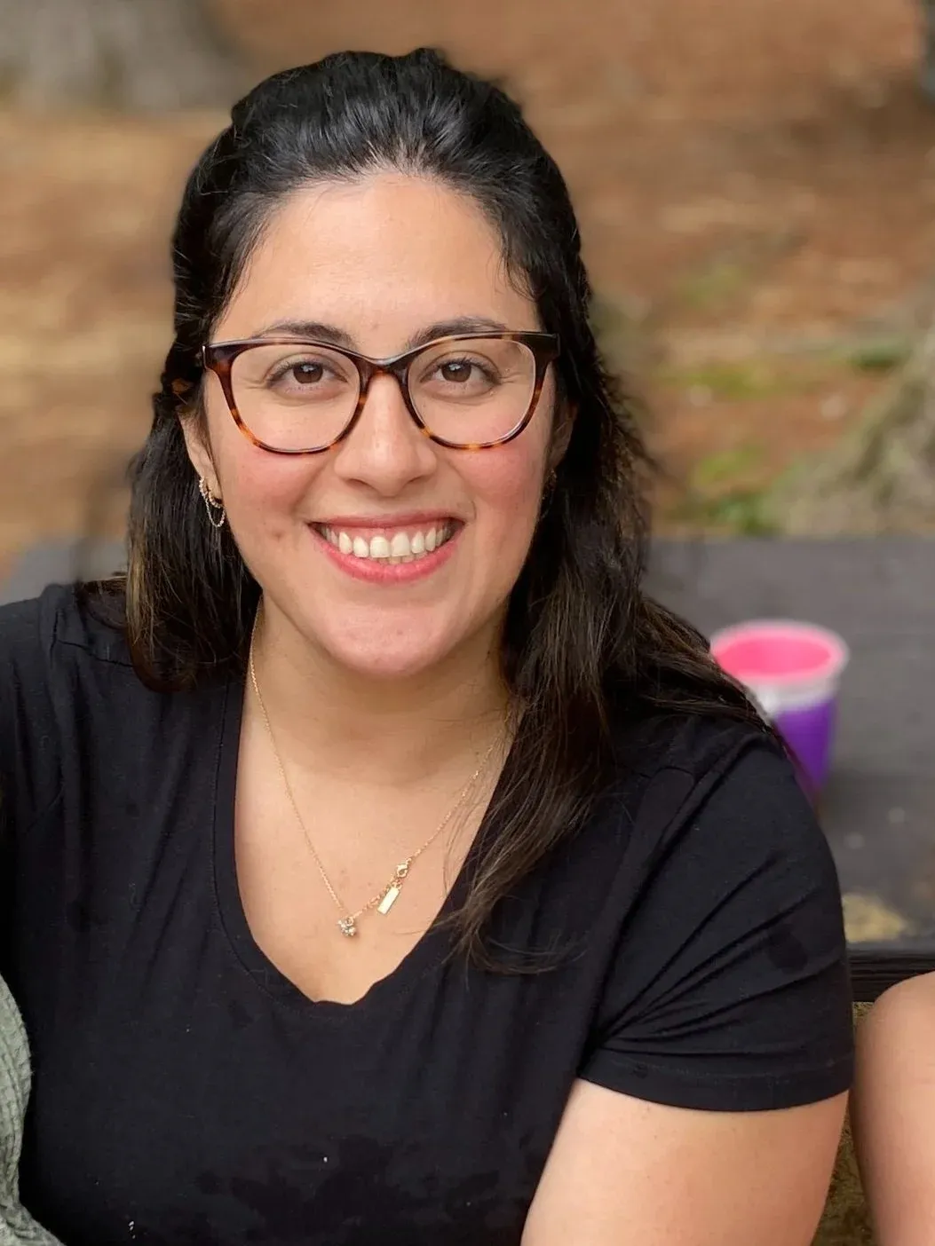 A woman with dark hair wearing glasses and jewelry smiling at the camera, sitting outside with a blurred background.
