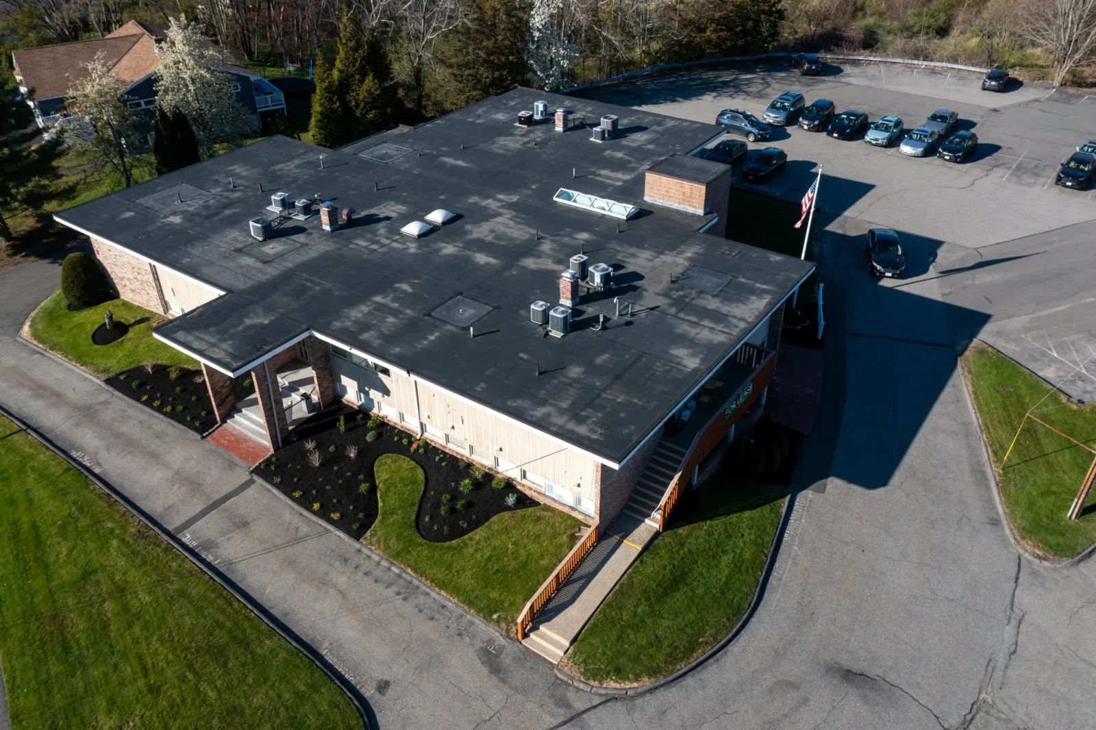 Aerial view of a one-story commercial building with a flat roof, surrounded by a parking lot with several cars, landscaped areas, and green lawns.