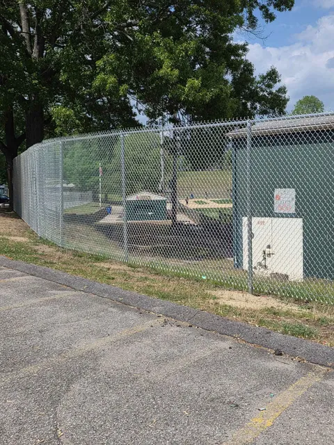 Chain-link fenced utility area beside a parking lot under trees on a sunny day