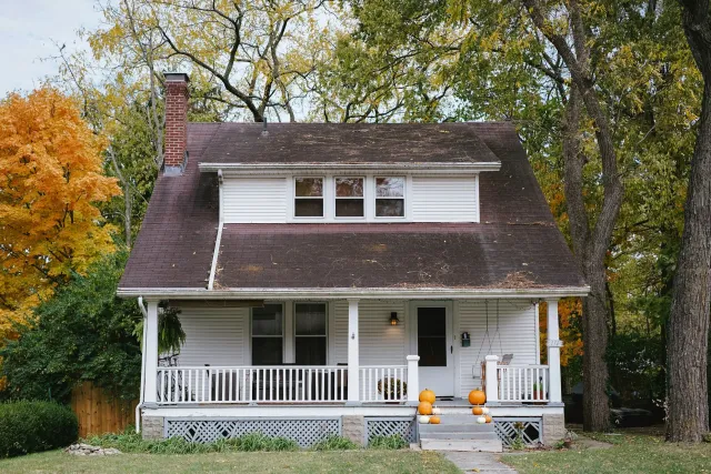 A two-story house with white siding, a front porch adorned with pumpkins, and surrounded by autumn trees with falling leaves.
