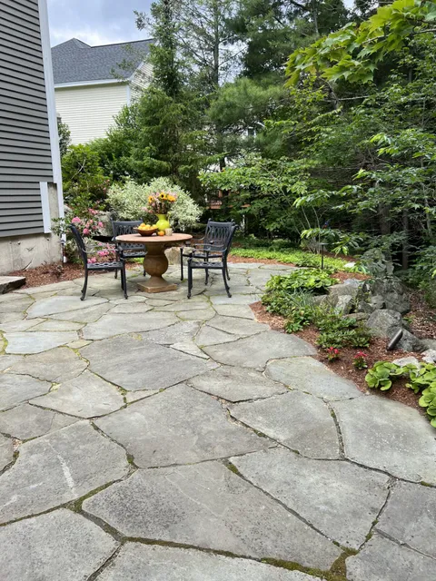 A serene garden patio with a stone floor, table, chairs, and lush greenery. Bright flowers and fruit centerpiece enhance the peaceful setting.