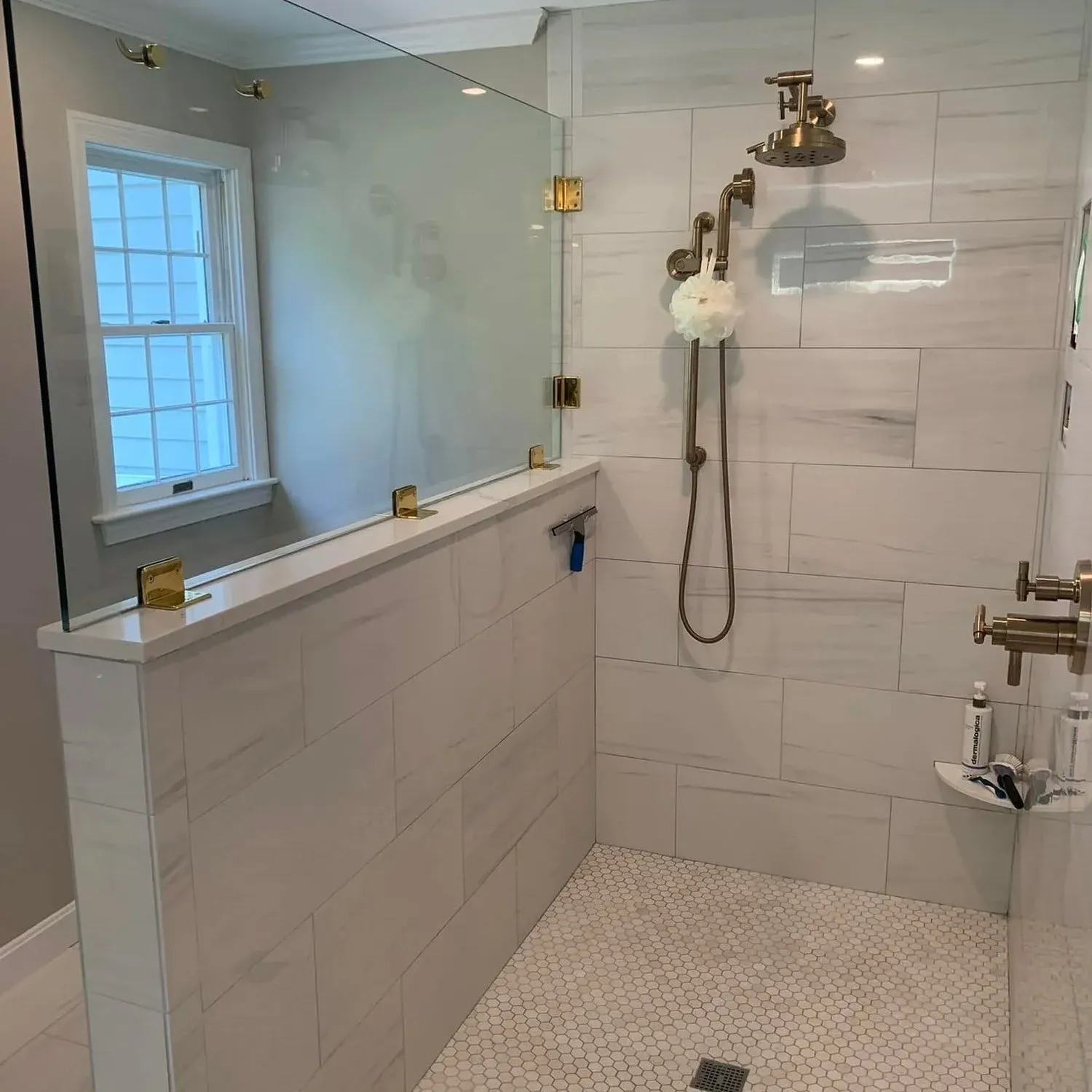 Modern bathroom shower area with a glass partition, white tiled walls, a window, and a shower fixture with a handheld showerhead. There are toiletries on a small shelf and a loofah hanging from the shower fixture.