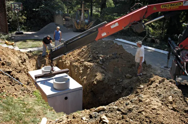 Construction workers using an excavator to install a large concrete tank in a dirt excavation site