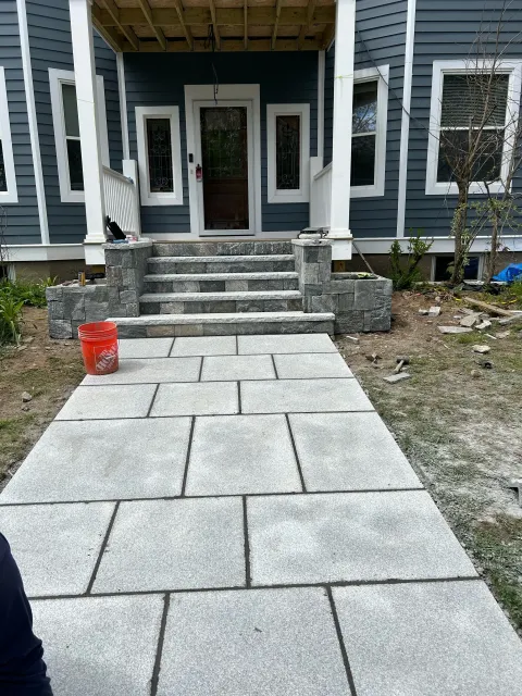 Front porch of a house with stone steps leading up to the door, blue siding, and a partially constructed wooden roof cover. Paving stones are being installed on the walkway in front of the porch.