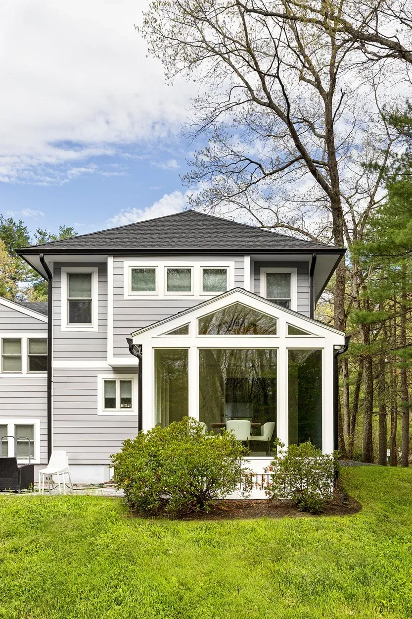 Back view of a modern gray two-story house with a sunroom and a well-maintained lawn surrounded by trees.