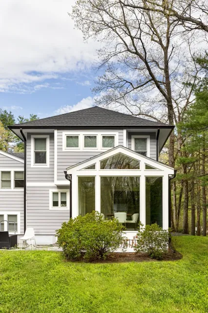 Back view of a modern gray two-story house with a sunroom and a well-maintained lawn surrounded by trees.
