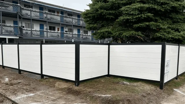 Photo of a white privacy fence with black framing around a tree in front of an apartment building with balconies.