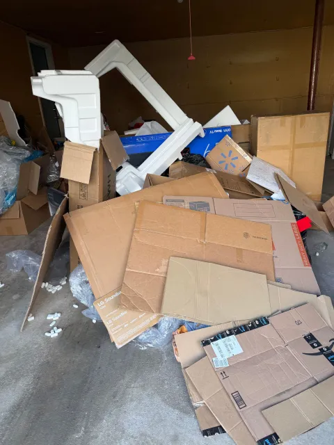 Overflowing cardboard boxes and debris piled in a dim storage room, with a white frame leaning against the wall.