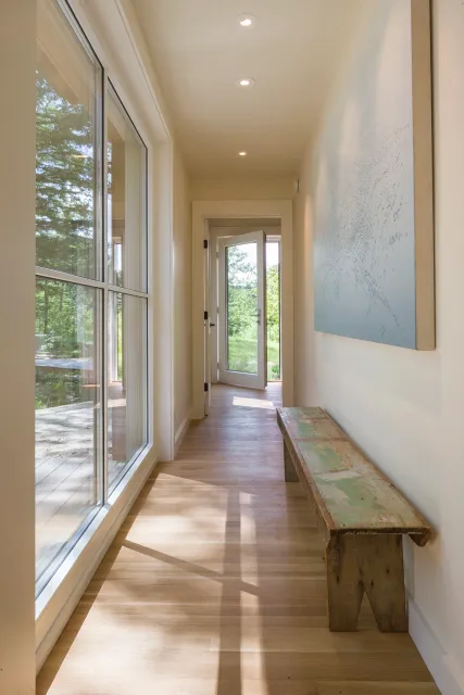 A bright hallway with a wooden bench on the right, a large window on the left revealing trees outside, and a door at the end leading to an outdoor area during the daytime.