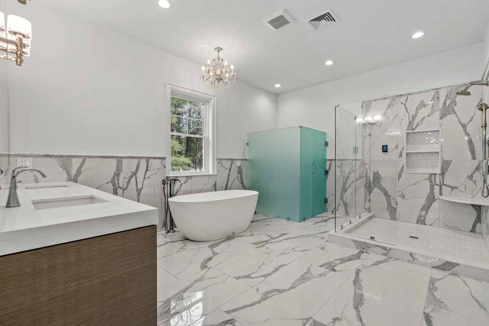 Modern bathroom with marble tiles, a freestanding bathtub, a glass shower enclosure, a double vanity, a window, and a chandelier.