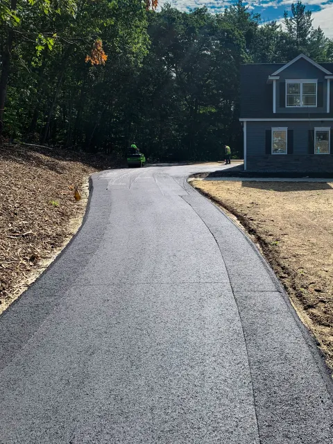 A newly paved asphalt driveway curves towards houses and a wooded area, with construction workers and equipment present.