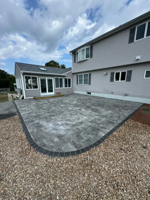 Backyard with newly installed paved patio and gravel border, connected to a house with siding and multiple windows.