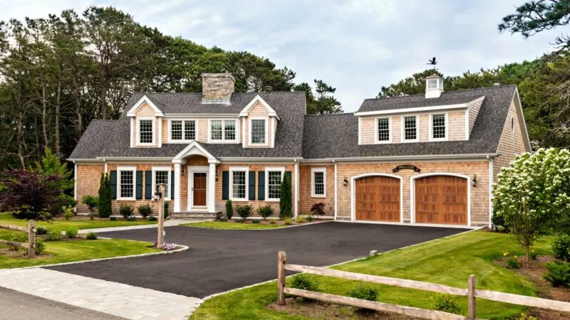 Spacious Cape Shingle Style house with tan shingles, white trim, dark gray roof, trees – McPhee Associates of Cape Cod.