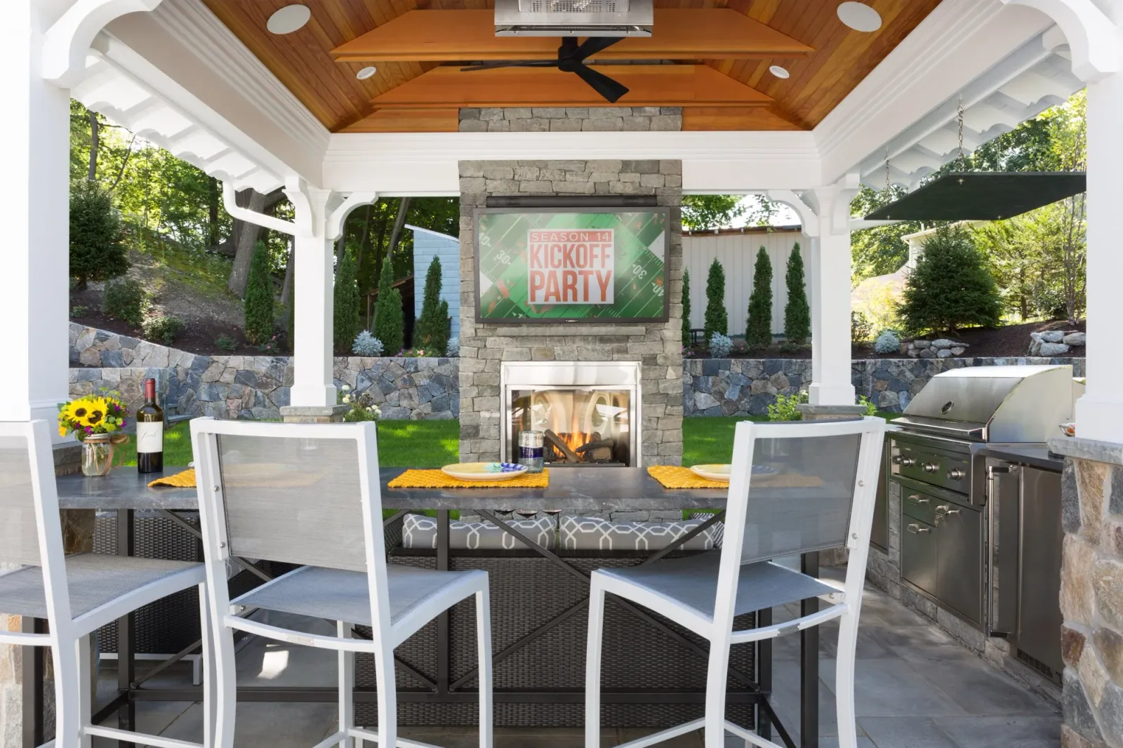 Outdoor patio with a stone fireplace, mounted TV showing a football kickoff party graphic, dining table with yellow placemats, wine bottle with sunflower arrangement, and a stainless steel grill, under a wooden ceiling with a ceiling fan.