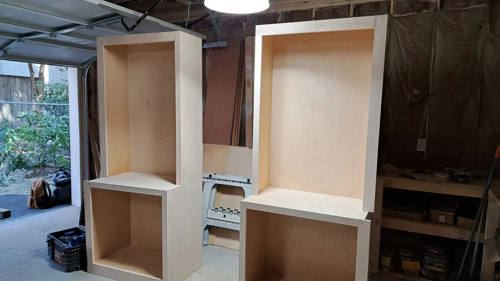 Two unfinished wooden cabinets with open shelves in a workshop, one white and one natural wood.