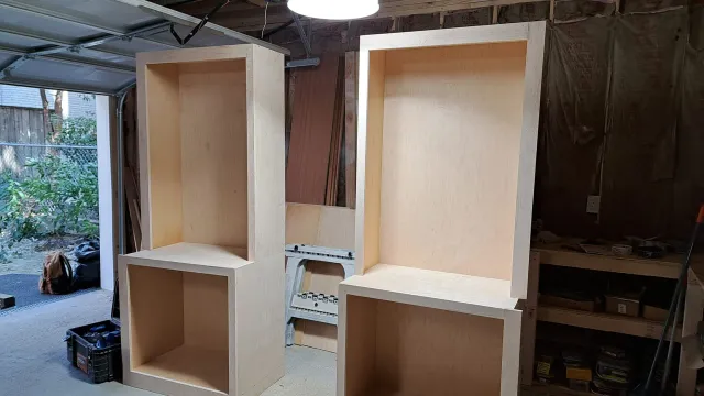Two unfinished wooden cabinets with open shelves in a workshop, one white and one natural wood.