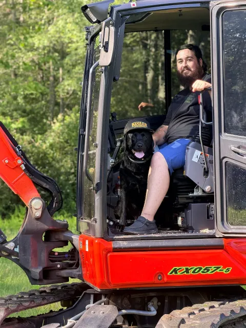 Man in a red excavator cab with a black dog, outdoors by a wooded area
