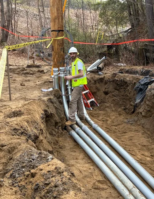 A construction worker wearing a white hard hat and yellow safety vest is standing next to large pipes in a dug trench, with construction materials and warning tape around the site.