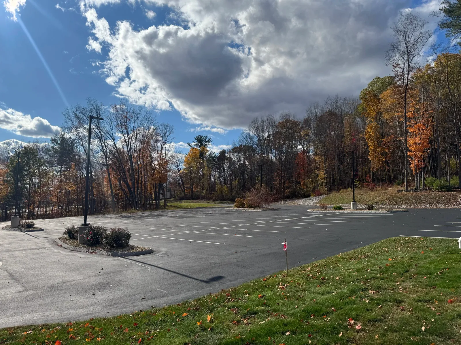 Empty parking lot surrounded by trees with autumn leaves, under a partly cloudy sky with the sun shining.