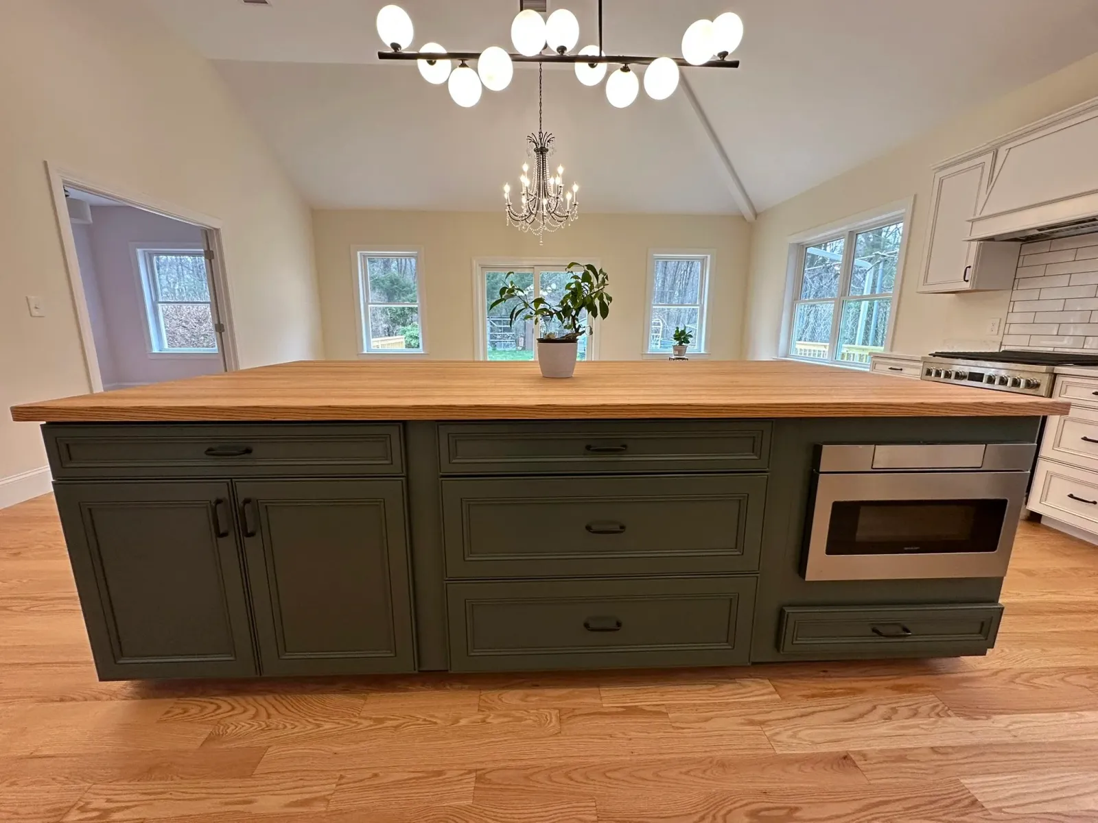 A modern kitchen with a large green island featuring drawers, a built-in microwave, and a wooden countertop. In the background, there are multiple windows, a chandelier and a ceiling light fixture, and a potted plant on the island.