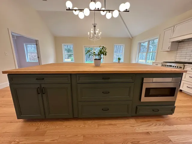 A modern kitchen with a large green island featuring drawers, a built-in microwave, and a wooden countertop. In the background, there are multiple windows, a chandelier and a ceiling light fixture, and a potted plant on the island.
