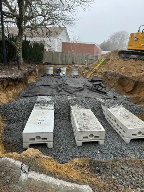 Construction site: excavation with rectangular concrete structures on gravel, black fabric, and a yellow hose.