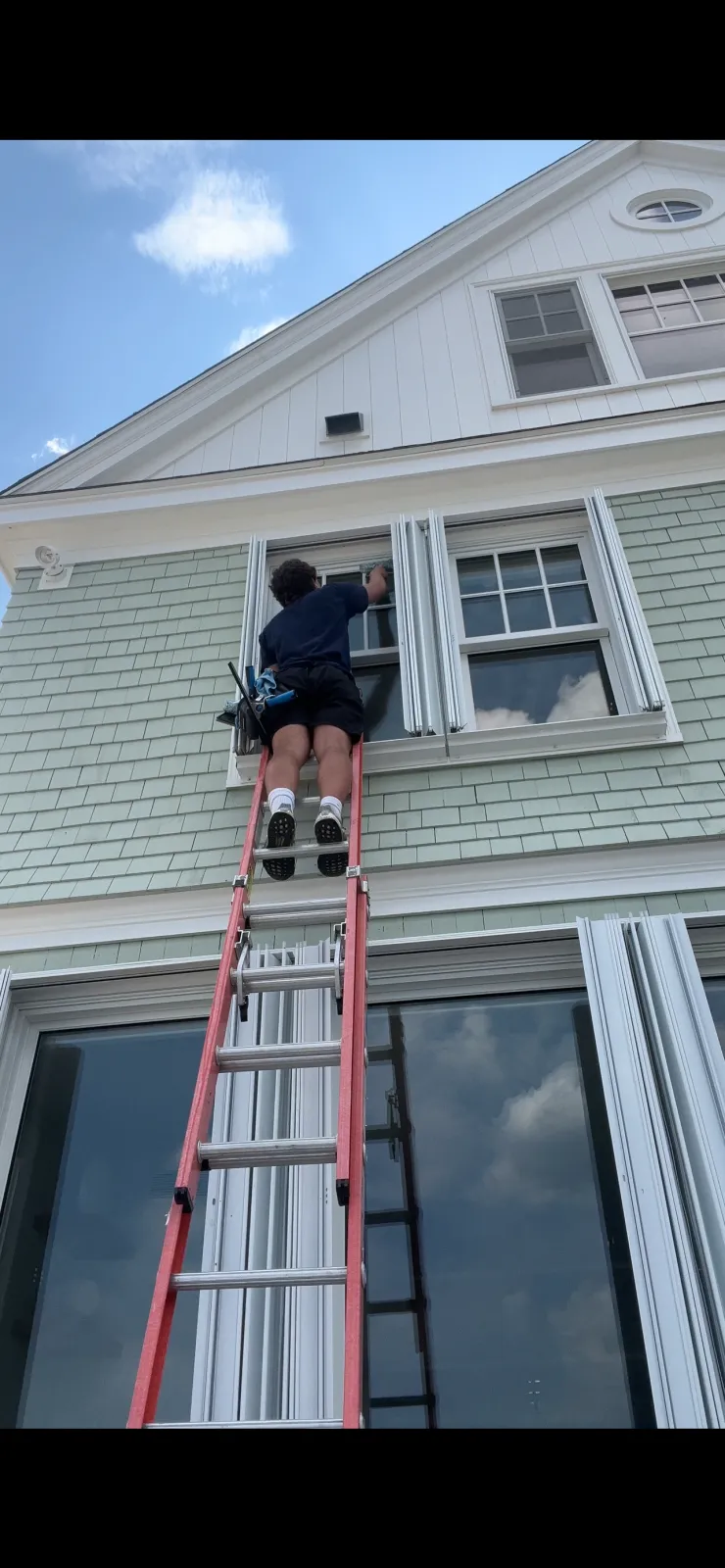 A person standing on a ladder cleaning or repairing the exterior window of a house with light green siding and white trim. The house has multiple windows and a gabled roof under a partly cloudy sky.