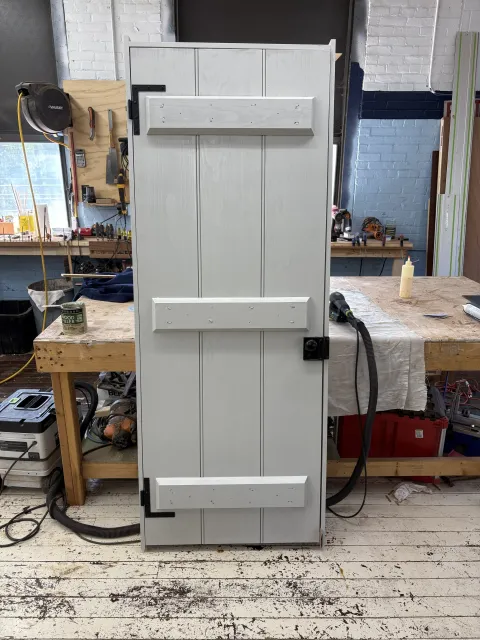 A woodworking workshop with a white wooden cabinet in the center, tools hanging on a board, a workbench with various tools and supplies, and a stained wooden floor.