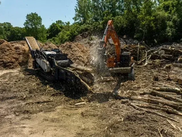 Excavator feeding logs into a wood chipper at a lumberyard, creating wood chips.