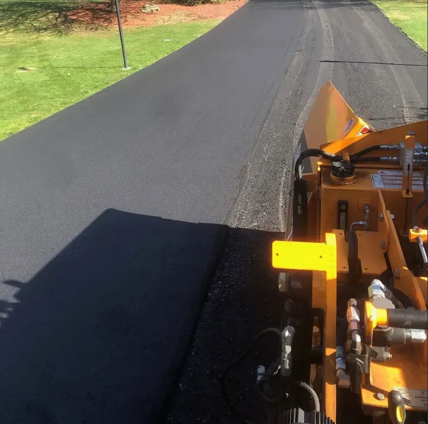 A view from the driver's seat of a yellow asphalt paving machine laying new blacktop on a driveway beside a green lawn.