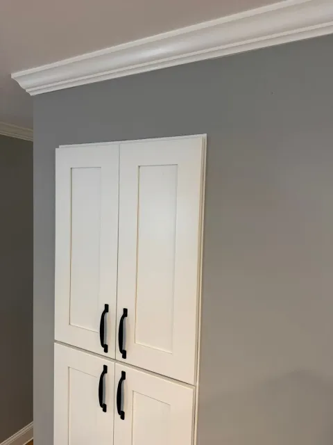 White wall cabinet with black handles mounted on a gray interior wall, beneath crown molding