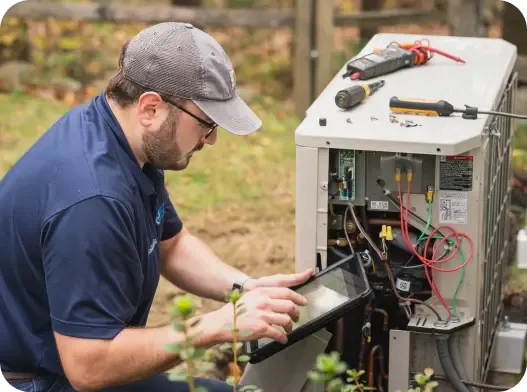 A technician wearing a cap and glasses uses a tablet while inspecting and working on an outdoor HVAC unit with tools placed on top.
