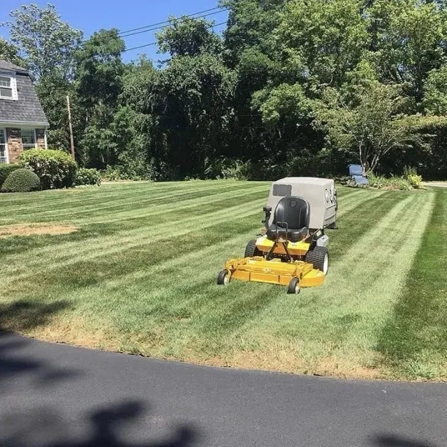 A yellow zero-turn lawn mower parked on a freshly mown lawn with distinct grass stripes in front of a house.