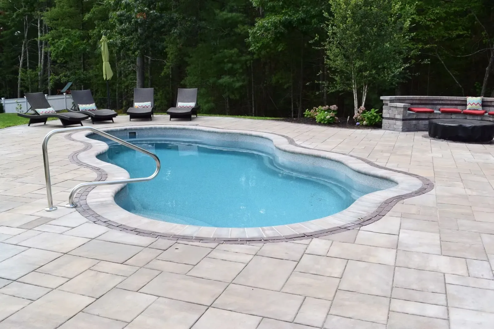 Outdoor pool with curved edges, surrounded by paving stones. Lounge chairs and trees in the background.