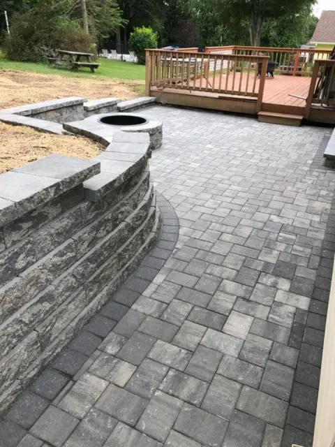 A backyard patio with gray interlocking brick pavers, a stacked stone retaining wall, a circular fire pit, and a wooden deck with steps leading down to the patio. A picnic table and trees are visible in the background.