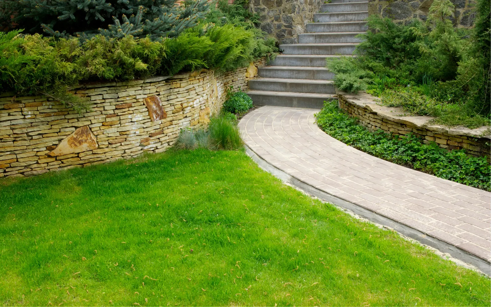 Garden path with curved brick walkway, stone steps, stone retaining walls, and green shrubs and grass.