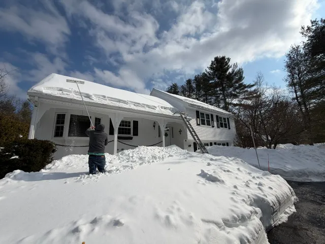 Snow-covered house with a person shoveling the front path under a cloudy winter sky