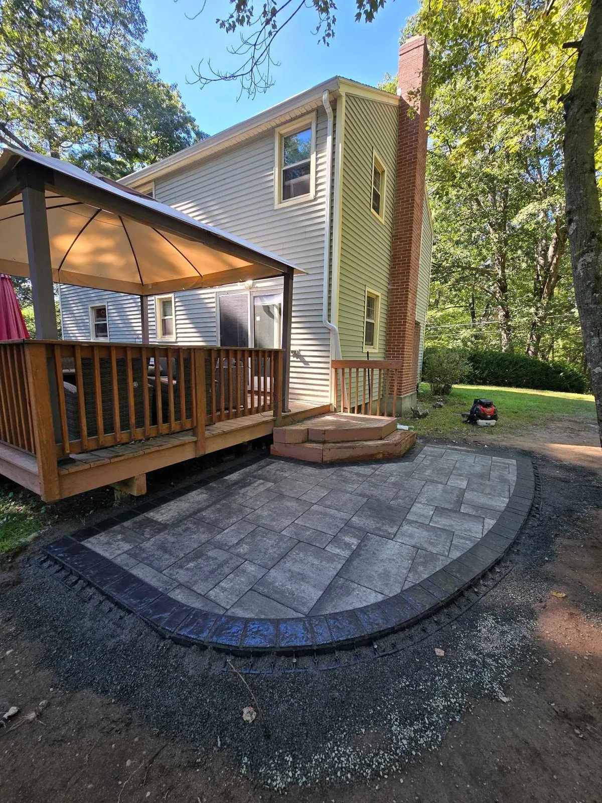 A backyard features a newly installed gray stone patio with a dark border, next to a wooden deck, gazebo, and house.