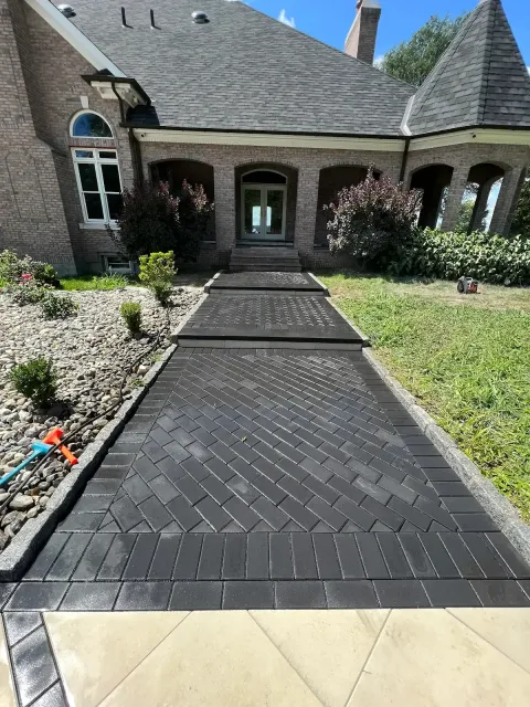 Newly installed black brick driveway leading to the front door of a brick house with landscaped garden on each side under a blue sky.