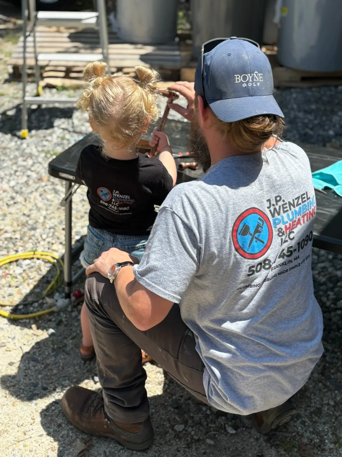 Man and young girl working together on plumbing outside, man wearing a gray shirt and cap with company's logo, girl with blonde hair in bun wearing a black shirt, sunlight, gravel ground.