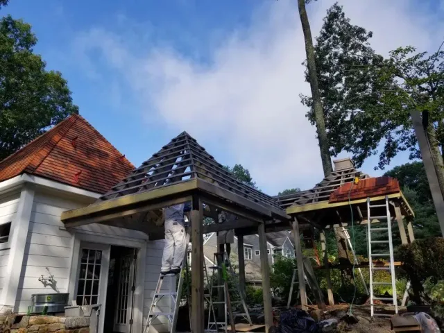 Construction of a gazebo addition to a white house with a red tile roof, ladders, and scaffolding under a cloudy sky.