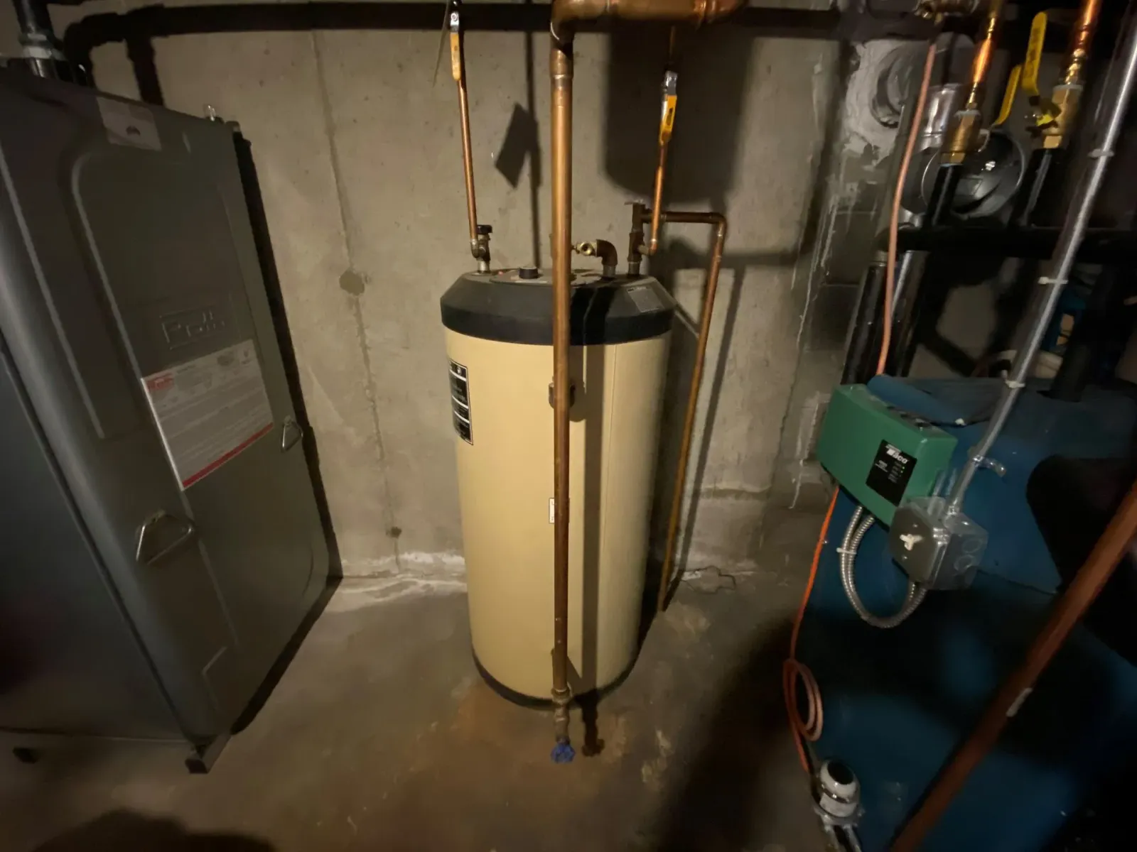 A beige water heater tank with metal pipes is installed in a dimly lit basement with concrete walls and floor, typical of homes in Norfolk and Suffolk County, MA. Equipment and machinery are visible nearby.